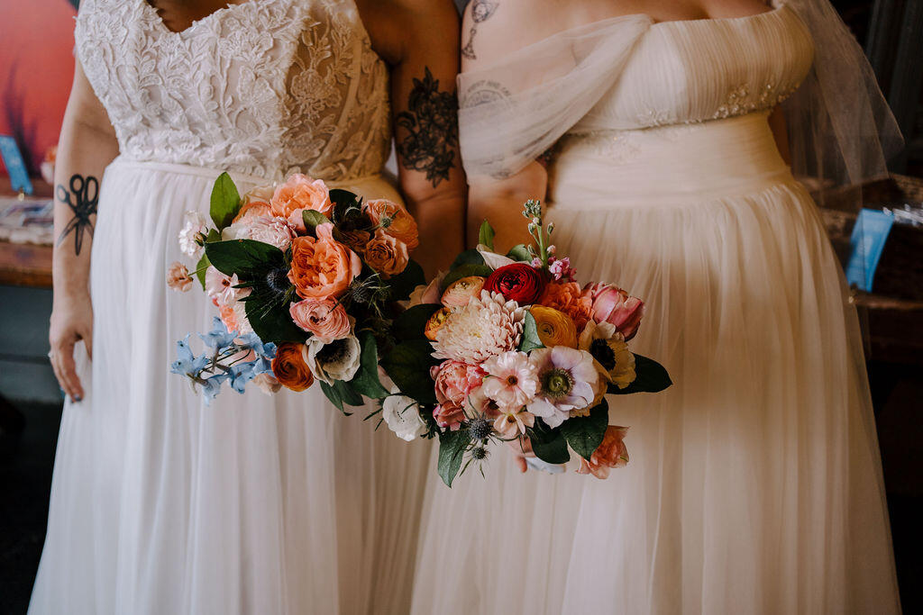 A full body shot of two woman wearing wedding dresses and holding bridal bouquets.