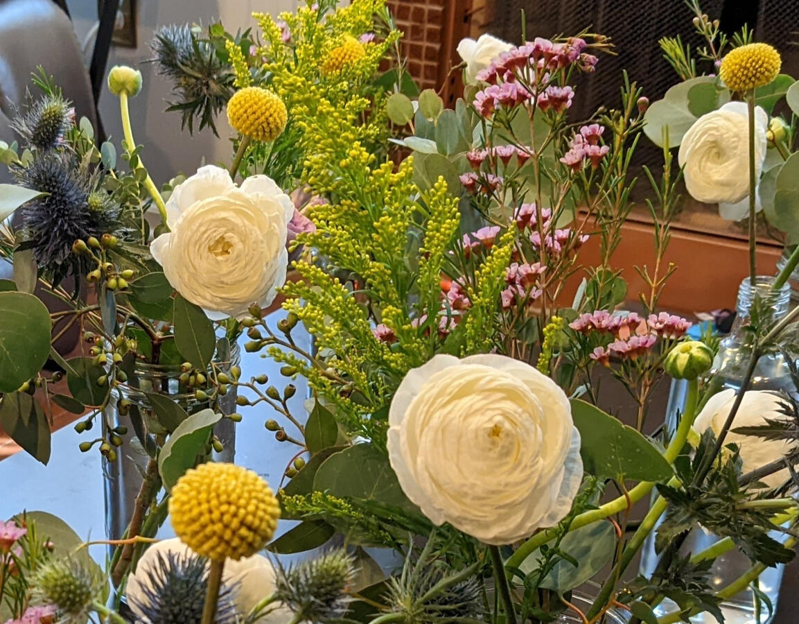 A table with various vases, filled with white ranunculus, thistle, wax flower, and small yellow flowers.