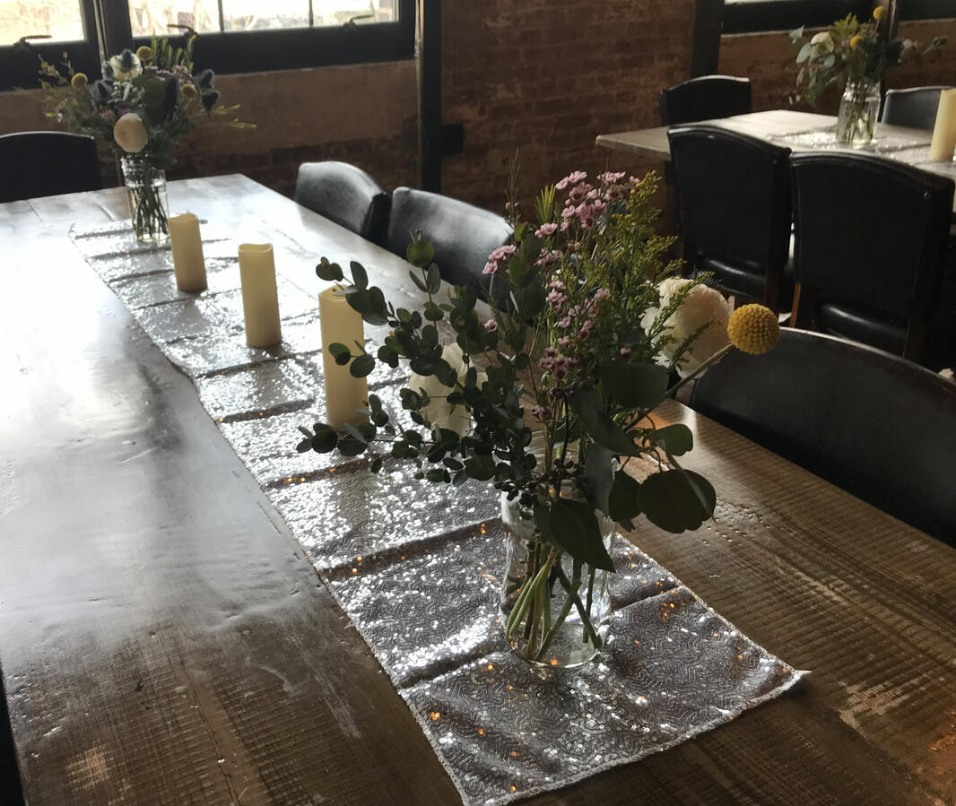 A long table with a silver sequined tablecloth and two large arrangements in glass vases.