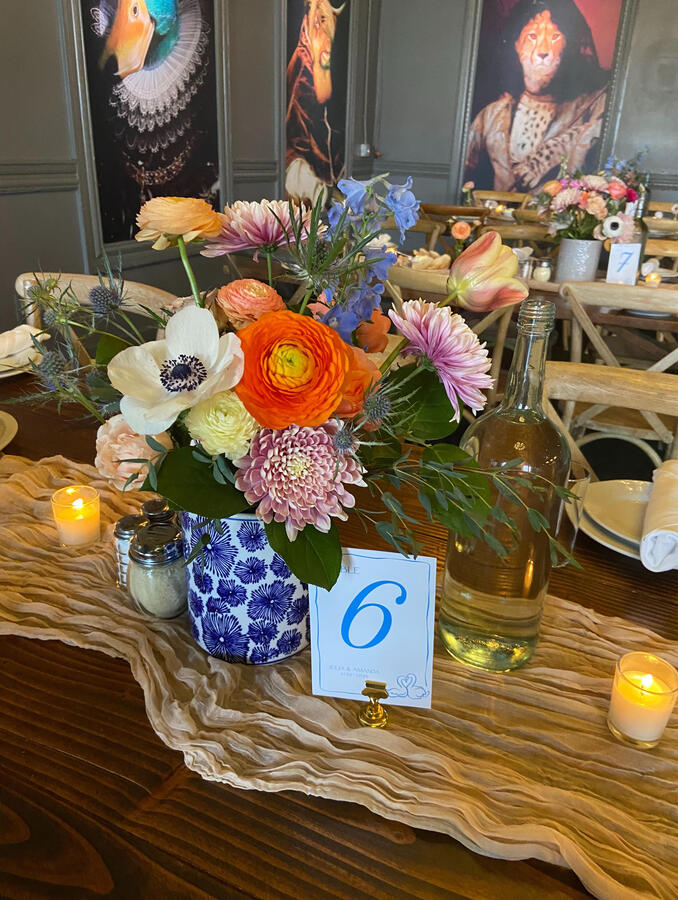 A long table with a gauze tablecloth, a large floral arrangement with several smaller bud vases, a votive candle and a glass water bottle. There's a table sign with the number six on it.