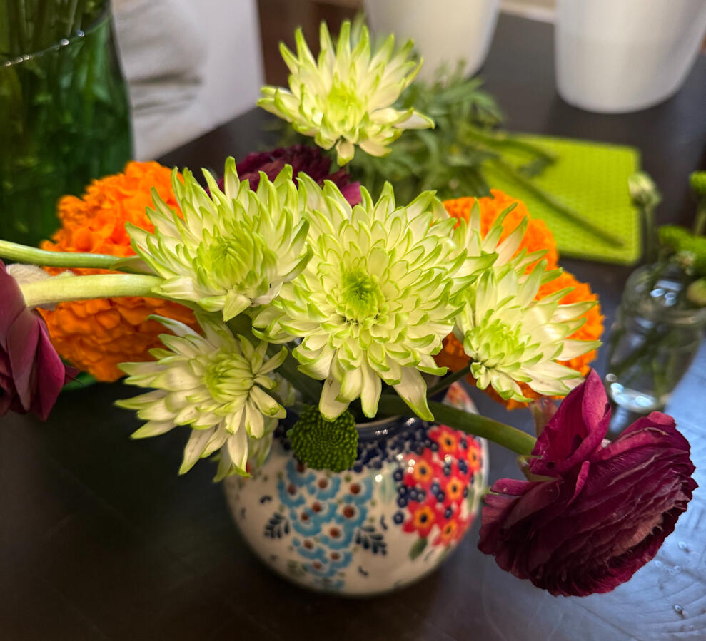 A small porcelain vase with green mums, orange marigolds and deep purple ranunculus.