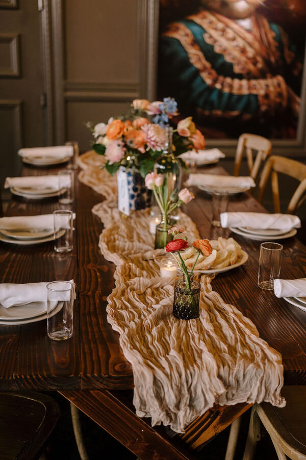 A long table with a gauze tablecloth, a large floral arrangement with several smaller bud vases, and votive candles.