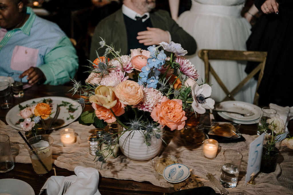 A long table with a gauze tablecloth, a large floral arrangement with several smaller bud vases, and votive candles.