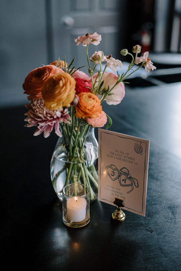A glass vase holding ranunculus blooms and mums, next to a lit votive candle and a table sign that reads "To all the love stories that came before us, thank you for showing us the way."