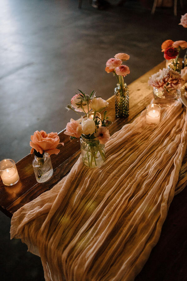 A table with a gauze tablecloth, votive candles, and several small bud vases with peach and pink flowers.