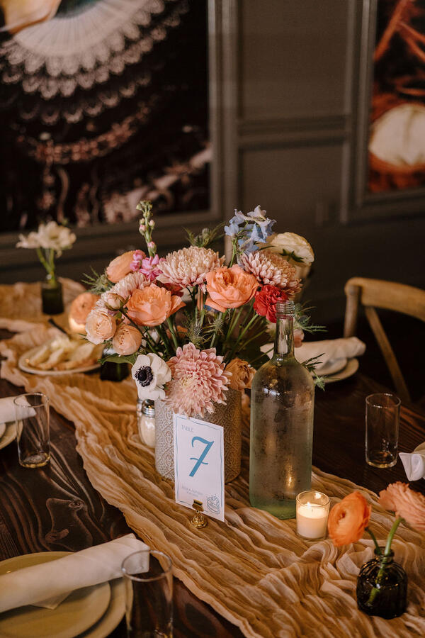 A long table with a gauze tablecloth, a large floral arrangement with several smaller bud vases, a votive candle and a glass water bottle. There's a table sign with the number seven on it.